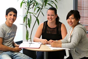 Three people at a table who look ready to learn English language skills with notepads and out looking at camera.